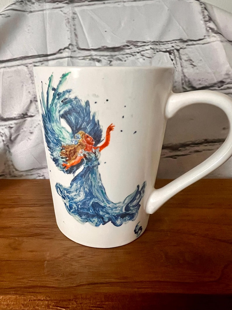 May include: White ceramic mug with a hand-painted design of a woman with blue hair and a blue dress, surrounded by blue and green water-like swirls.