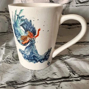 May include: White ceramic mug with a blue and green watercolor design of a woman with flowing hair and a blue dress. The woman is surrounded by water.