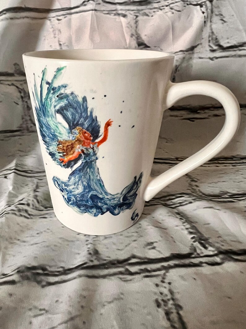 May include: White ceramic mug with a blue and green watercolor design of a woman with flowing hair and a blue dress. The woman is surrounded by water.
