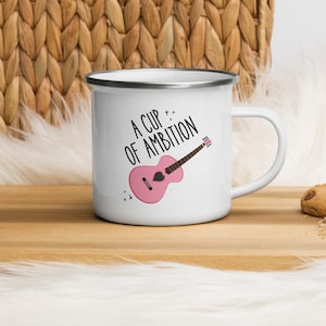 以下が含まれることがあります： ピンクのギターと「A Cup of Ambition」というテキストの白黒のグラフィックが描かれた白いエナメルマグカップです。
