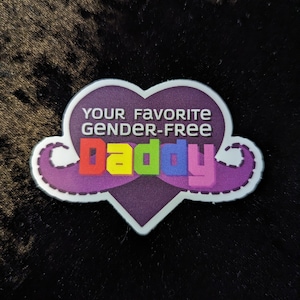 Peut inclure: Autocollant en forme de cœur violet avec le mot "Daddy" en arc-en-ciel et le texte "Your Favorite Gender-Free" au-dessus.