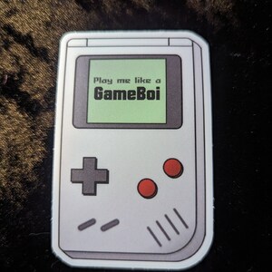 Peut inclure: Autocollant représentant une console de jeu portable rétro. L'appareil blanc affiche le texte "Play me like a GameBoi" sur un écran vert. Il comporte une croix directionnelle grise, des boutons rouges et des fentes de haut-parleur.