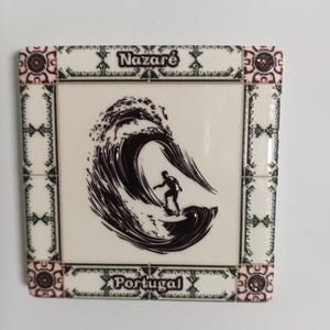 Pode incluir: Azulejo decorativo com um surfista a surfar uma onda. O azulejo tem as palavras "Nazaré" e "Portugal" numa moldura decorativa com detalhes em vermelho e verde. O surfista e a onda são pretos.