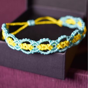 May include: A handmade macrame bracelet in a box. The bracelet features a repeating pattern of light blue and yellow knots. The adjustable closure is yellow. The bracelet is displayed on a dark purple surface.