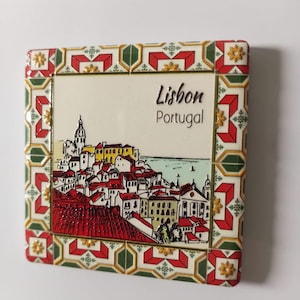 Może przedstawiać: Kwadratowy magnes ceramiczny z wizerunkiem Lizbony w Portugalii. Obraz przedstawia budynki z czerwonymi dachami, kościół i napis "Lisbon Portugal". Obramowanie ma dekoracyjny wzór w kolorze czerwonym, zielonym i złotym.