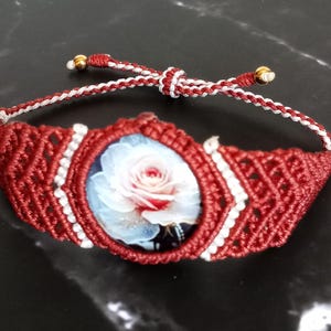 May include: A macrame bracelet featuring a central oval cabochon with a printed image of a white and pink rose. The bracelet is crafted from deep red and white cord in a geometric pattern, finished with adjustable sliding knots and gold beads.