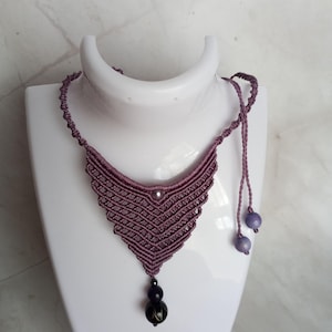 May include: A mauve macrame necklace featuring a triangular woven pattern with a central bead, a dangling pendant composed of two dark spherical beads, and two additional light purple beads on the adjustable cord ends.
