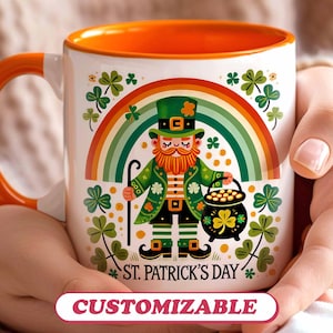 May include: A white ceramic mug with an orange rim features a cartoon illustration of a leprechaun holding a pot of gold. The leprechaun is standing in front of a rainbow with shamrocks and the text "St. Patrick's Day". The mug is customizable.