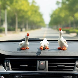 May include: Three small white duck figurines with orange beaks and feet are positioned on a black car dashboard. The left and right ducks are standing with their heads tilted upward, while the center duck is positioned in a downward-facing pose.