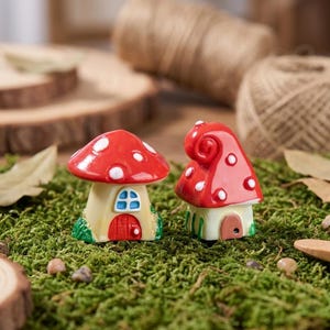 May include: Two miniature mushroom-shaped houses made of ceramic. The left house has a wide red cap with white spots, a cream-colored base, a blue window, and a red door. The right house has a tall, curled red cap with white spots and a brown door.