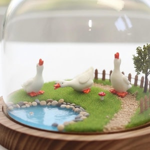 May include: A miniature diorama enclosed in a glass dome on a wooden base featuring three white geese with orange beaks and feet. The scene includes a small blue pond with pebbles, a green grassy area, a gravel path, a tiny red mushroom, and a tree.