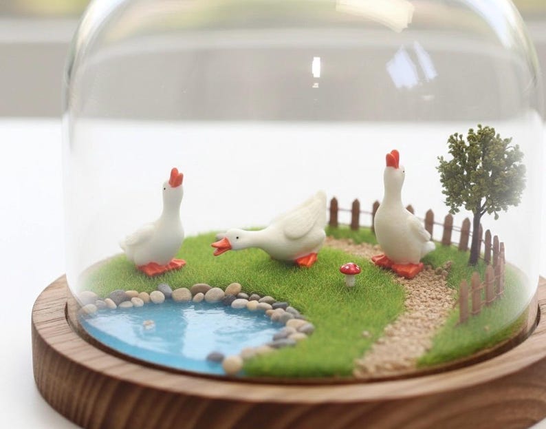 May include: A miniature diorama enclosed in a glass dome on a wooden base featuring three white geese with orange beaks and feet. The scene includes a small blue pond with pebbles, a green grassy area, a gravel path, a tiny red mushroom, and a tree.