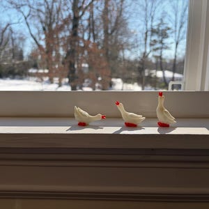 May include: Three small white goose figurines with bright red beaks and feet. The first goose is positioned with its head lowered, the second is standing in a neutral pose, and the third is standing with its head tilted upward toward the sky.