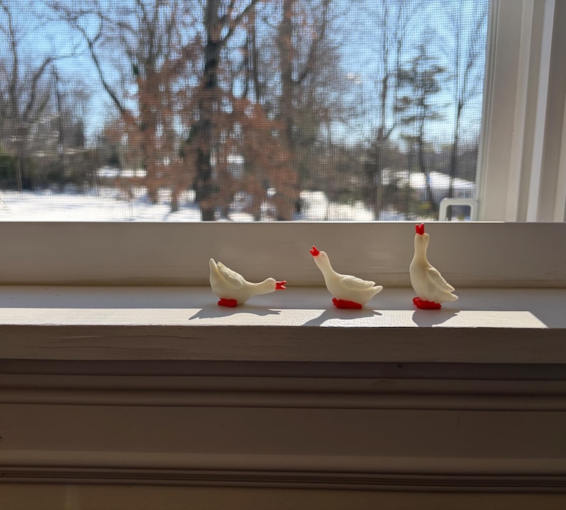 May include: Three small white goose figurines with bright red beaks and feet. The first goose is positioned with its head lowered, the second is standing in a neutral pose, and the third is standing with its head tilted upward toward the sky.