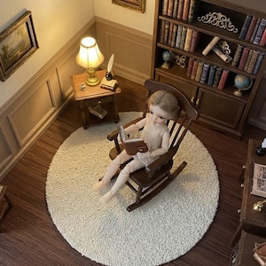 Può includere: Una scena in miniatura di una casa delle bambole con una piccola bambola articolata seduta su una sedia a dondolo in legno scuro sopra un tappeto rotondo bianco. La stanza include una libreria in legno piena di libri minuscoli, un tavolo e una penna.