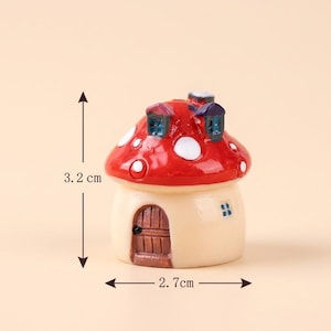 May include: A miniature mushroom-shaped house figurine with a glossy red cap featuring white spots and two small dormer windows. The base is cream-colored with a brown arched door and a small window. It measures 3.2 cm in height and 2.7 cm in width.