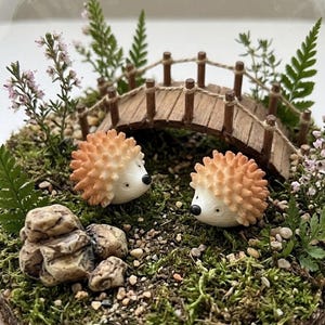May include: A miniature terrarium featuring two hedgehog figurines with orange and white quills, set amidst a mossy landscape with a small wooden bridge and decorative plants. The scene includes small rocks and delicate flowers.