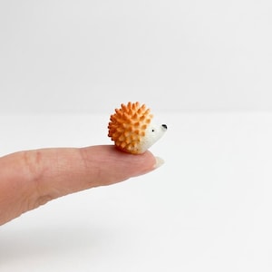Puede incluir: Una figura de erizo en miniatura con el lomo naranja texturizado y la cara blanca lisa. La pequeña figura animal presenta un hocico puntiagudo, una diminuta nariz negra y un único ojo negro visible, con una superficie espinosa detallada.