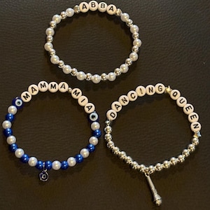 May include: Three beaded bracelets. One spells "ABBA" with white letter beads and silver beads. Another spells "MAMMA MIA" with white letter beads, blue and white beads, and a blue charm. The third spells "DANCING QUEEN" with white letter beads, silver beads, and a microphone charm.
