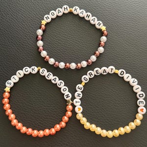 May include: Three beaded bracelets. One spells "NOAH KAHAN", another "STICK SEASON", and the third "THE GREAT DIVIDE". They have gold star and heart charms, and are made with pearl and coloured beads.