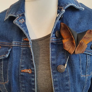 May include: A butterfly-shaped brooch with a paisley pattern in shades of orange, brown, and burgundy, pinned to a denim jacket. The brooch is attached with a silver pin. The jacket is a medium wash denim.