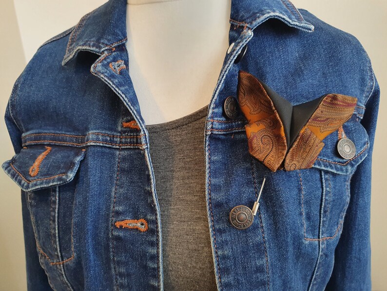May include: A butterfly-shaped brooch with a paisley pattern in shades of orange, brown, and burgundy, pinned to a denim jacket. The brooch is attached with a silver pin. The jacket is a medium wash denim.