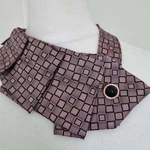 May include: A mauve ascot with a geometric square pattern and a black button with a gold-colored rim. The ascot is displayed on a white mannequin. The fabric is folded and buttoned.