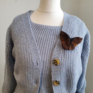 May include: A light blue knit cardigan with a matching sweater underneath. A brown and black butterfly brooch is pinned to the cardigan. The cardigan has three tortoise shell buttons.