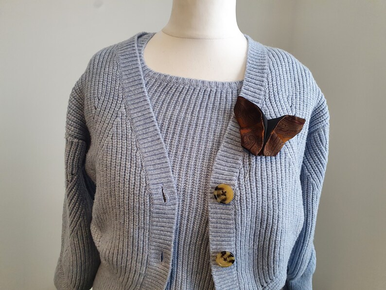May include: A light blue knit cardigan with a matching sweater underneath. A brown and black butterfly brooch is pinned to the cardigan. The cardigan has three tortoise shell buttons.