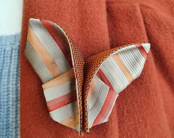 May include: A folded, striped fabric lapel flower in shades of orange, red, and tan, pinned to a rust-colored wool coat. A silver pin is visible below the flower, adding a touch of elegance to the attire.