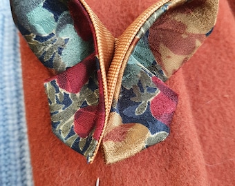 May include: A butterfly-shaped lapel pin with a floral pattern in shades of green, red, and gold. The pin has a gold center and a silver pin backing. The pin is set against a rust-colored fabric.