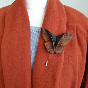 May include: A butterfly-shaped brooch with a patterned design in shades of brown, black, and orange is pinned to a rust-colored coat. The brooch is secured with a silver pin, adding a touch of elegance to the garment.