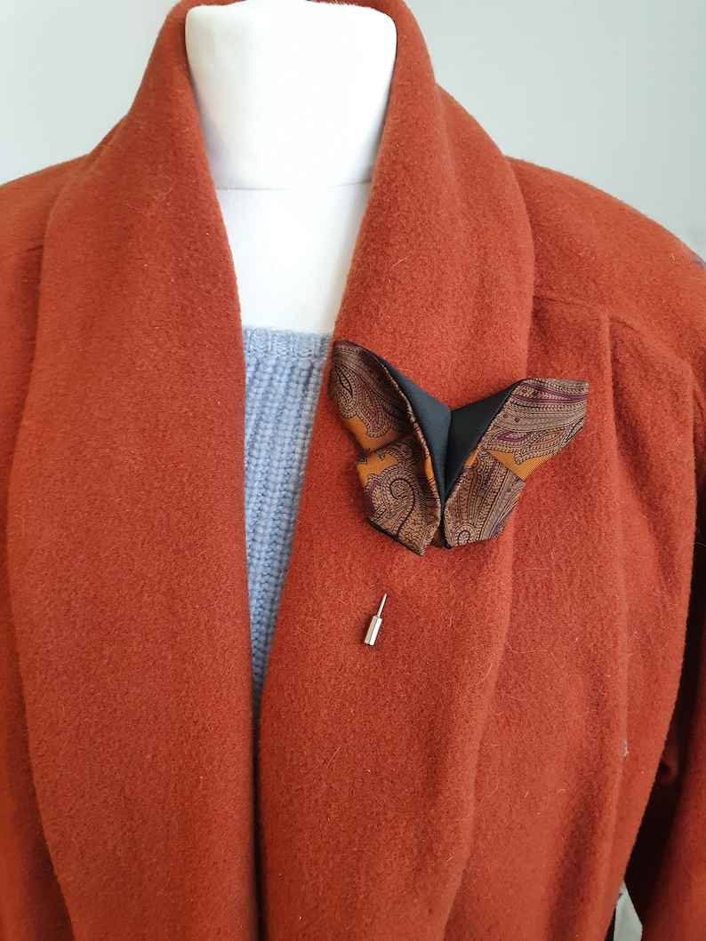 May include: A butterfly-shaped brooch with a patterned design in shades of brown, black, and orange is pinned to a rust-colored coat. The brooch is secured with a silver pin, adding a touch of elegance to the garment.