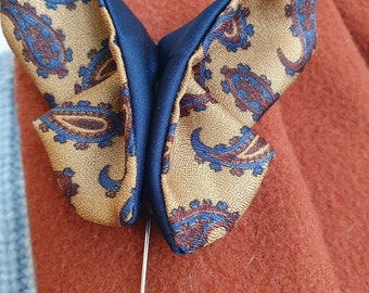 May include: A butterfly-shaped lapel pin. The pin features a gold fabric with a paisley pattern in blue and brown, contrasted by a dark blue center. A silver pin extends from the back.