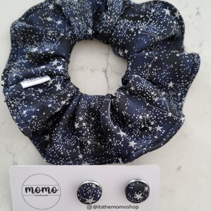 May include: A navy blue scrunchie and matching stud earrings with a white star pattern. The scrunchie is made of a soft fabric and is circular. The earrings are small, round, and have a silver border. The brand name "momo" is on the card.