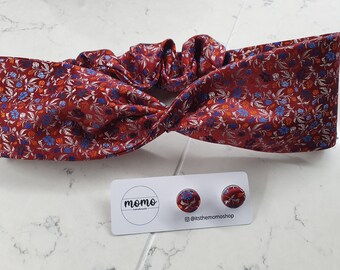 Puede incluir: Una diadema burdeos y pendientes a juego con un estampado floral en azul, naranja y plateado. La diadema está torcida en la parte superior. Los pendientes son redondos y tienen el mismo diseño floral.