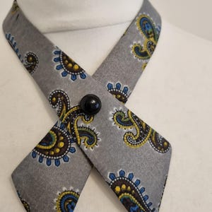 Könnte beinhalten: Ein graues Ascot mit einem Paisley-Muster in Gelb, Blau und Braun. Das Ascot wird mit einem schwarzen Knopf befestigt. Der Stoff ist um einen weißen Mannequinkragen gewickelt.