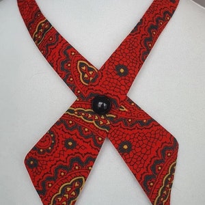 Könnte beinhalten: Eine rot gemusterte Krawatte mit einem schwarzen Knopfverschluss. Die Krawatte hat ein sich wiederholendes Paisley-ähnliches Design in Schwarz und Gold auf rotem Hintergrund. Die Krawatte wird auf einer weißen Schaufensterpuppe präsentiert.