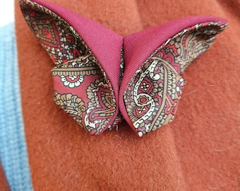 May include: A burgundy butterfly-shaped pocket square with a paisley pattern in red, brown, and cream. The pocket square is folded to resemble a butterfly and is paired with a silver lapel pin. The background is a rust-colored fabric.