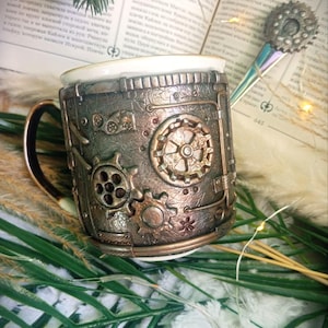 May include: Steampunk-themed mug featuring bronze and copper-toned gears and mechanical embellishments. The mug has a white interior and a gold handle. A decorative spoon with a gear-shaped top rests on a book in the background.