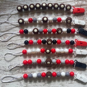 May include: Six baby pacifier clips with various bead designs. Each clip features a different color scheme, including red, white, gray, black, and brown beads. The clips have a loop for attaching to a pacifier and a plastic clip for securing to clothing.
