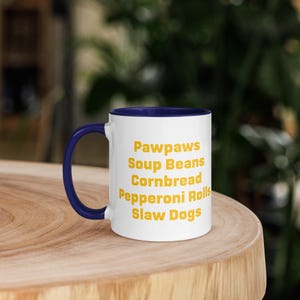 Pode incluir: Uma caneca de cerâmica branca com interior e alça azul-escura. O exterior apresenta texto amarelo listando as palavras Pawpaws, Soup Beans, Cornbread, Pepperoni Rolls e Slaw Dogs em uma pilha vertical. A caneca tem um formato cilíndrico.