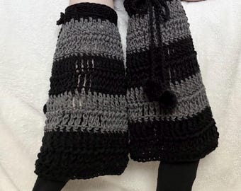 May include: A pair of crochet leg warmers featuring horizontal stripes in alternating black and dark gray yarn. The leg warmers have a textured, open-stitch pattern and include black drawstring ties at the top with small pom-pom accents.