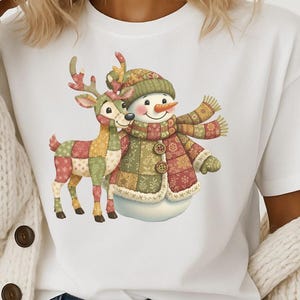 以下が含まれることがあります： 雪だるまとトナカイのイラストが描かれた白いTシャツ。雪だるまは緑の帽子とスカーフを身につけ、トナカイはパッチワークのデザインです。デザインは中央に配置され、陽気で心温まるイメージを作り出しています。