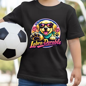 May include: Black t-shirt featuring a cartoon Labrador puppy wearing sunglasses and a bandana. The puppy is inside a circular design with hearts and the text "Labra-Dorable" in pink and yellow. A soccer ball is held in the image.