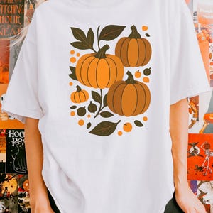 Può includere: T-shirt bianco con grafica a tema autunnale. Il design include tre zucche arancioni di varie dimensioni, foglie verde scuro e piccoli punti arancioni. La maglietta è in stile casual, perfetta per l'autunno.