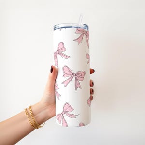 May include: White tumbler with a repeating pattern of pink bows outlined in black. The tumbler has a silver lid and a clear straw. The tumbler is held by a person with gold bracelets.
