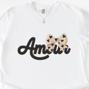 May include: White t-shirt with the word "Amour" in black script, a beige bow with black heart accents, and a silver necklace with a heart pendant. The shirt is a Comfort Colors brand.