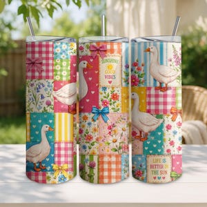 May include: Three cylindrical tumblers with straws featuring a patchwork design of white geese, colorful flowers, pink and yellow gingham patterns, and bows. Text on the tumblers reads "SUNSHINE GOOD VIBES" and "LIFE IS BETTER IN THE SUN" with small hearts.