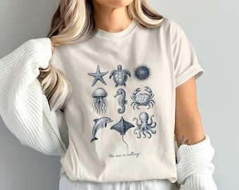 May include: Cream-colored t-shirt featuring a collection of marine life illustrations in dark blue. The design includes a starfish, turtle, jellyfish, seahorse, crab, dolphin, stingray, and octopus. The text "the sea is calling" is printed below the illustrations.